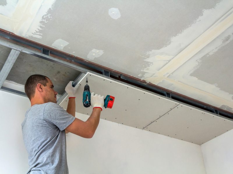 Construction worker assemble a suspended ceiling with drywall and fixing the drywall to the ceiling metal frame with screwdriver.